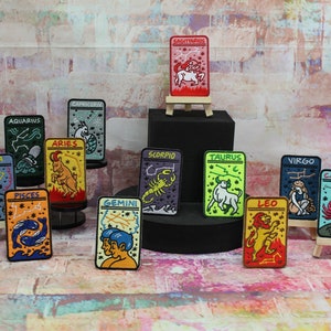 May include: Twelve colourful embroidered iron-on patches featuring the astrological signs of the zodiac. The patches are arranged on a black surface and a wooden easel. The signs include Aries, Taurus, Gemini, Cancer, Leo, Virgo, Libra, Scorpio, Sagittarius, Capricorn, Aquarius, and Pisces.