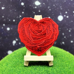 May include: A heart-shaped, red embroidered rose patch. The patch is displayed on a small wooden easel against a backdrop of a starry night. The rose is detailed with black thread, showcasing the petals.
