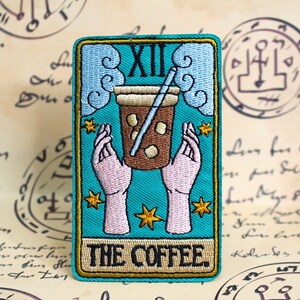 May include: A teal blue tarot card with gold trim and a brown coffee cup with a straw. The card is labelled "XII THE COFFEE".