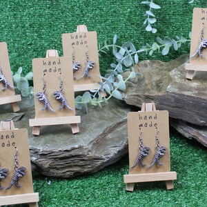 May include: Five pairs of silver dinosaur skeleton earrings displayed on small wooden easels. Each pair of earrings is labelled "hand made".