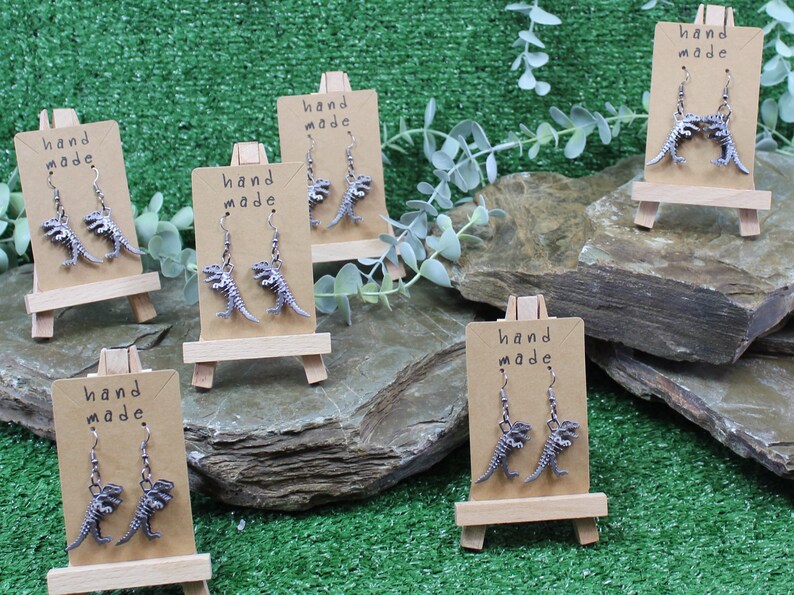 May include: Five pairs of silver dinosaur skeleton earrings displayed on small wooden easels. Each pair of earrings is labelled "hand made".