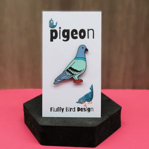 May include: A blue and green enamel pin of a pigeon with red eyes. The pin is on a white card with the word "pigeon" in black text and the words "Fluffy Bird Design" in black text.