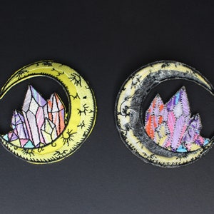 May include: Two embroidered iron-on patches featuring a crescent moon with a cluster of colorful crystals inside. One patch has a yellow moon and the other has a black moon.