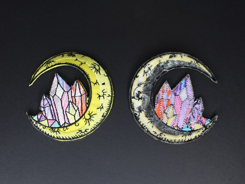 May include: Two embroidered iron-on patches featuring a crescent moon with a cluster of colorful crystals inside. One patch has a yellow moon and the other has a black moon.