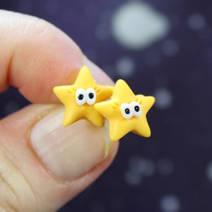 Close-up of a single pair of star earrings held between fingers, showing its size and detailing. -  Brighten your everyday look with these yellow star stud earrings, a tiny pop of sunshine that catches the light with every turn. Their cheerful star shape and warm yellow hue add a sweet, celestial sparkle that feels playful yet polished.