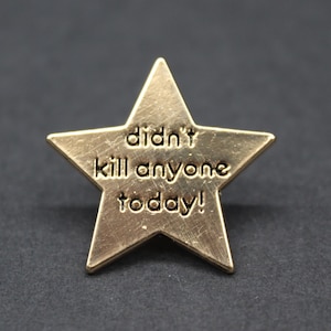 A gold star-shaped enamel pin with the words “didn’t kill anyone today!” engraved on the front. The pin has a brushed metallic finish and sits against a dark grey background, creating a playful but stylish contrast. A humorous novelty gift for adults who appreciate sarcasm and self-aware humour, ideal for workmates, friends, or anyone needing a lighthearted reminder that they made it through the day.