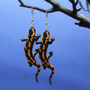 May include: A pair of salamander-shaped dangle earrings. The earrings are black with orange and yellow patterns, hanging from silver-tone hooks. The background is a solid blue colour.