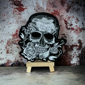 May include: Black and white embroidered skull with floral details, displayed on a small wooden easel.