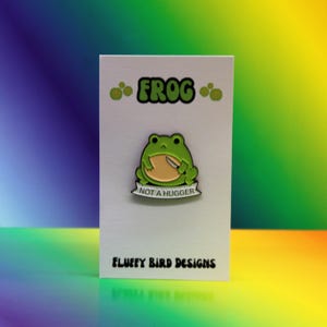 May include: A green frog enamel pin with a tan belly and a silver knife, displayed on a white card. The card reads "FROG" above the pin and "NOT A HUGGER" below. The card also says "FLUFFY BIRD DESIGNS".