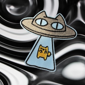 May include: An embroidered patch featuring a cat in a flying saucer. The saucer is light brown with a black outline. The cat is yellow with a black outline and is looking out of the saucer. The patch is on a black and white background.