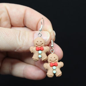 K&ouml;nnte beinhalten: Ein Paar Lebkuchenmann-Ohrringe mit roten Schleifen und blauen und wei&szlig;en Zuckergussdetails. Die Ohrringe sind aus Polymer Clay gefertigt und haben silberne Haken.