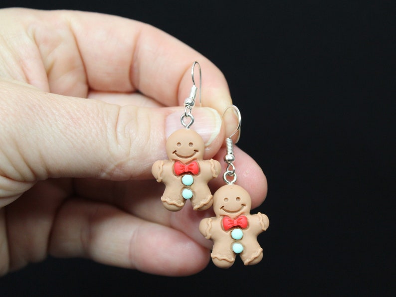 K&ouml;nnte beinhalten: Ein Paar Lebkuchenmann-Ohrringe mit roten Schleifen und blauen und wei&szlig;en Zuckergussdetails. Die Ohrringe sind aus Polymer Clay gefertigt und haben silberne Haken.