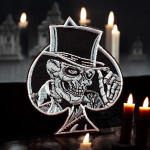 May include: A black and white embroidered patch depicting a skull wearing a top hat and holding a playing card. The card is a spade and the skull is looking to the right. The patch is set against a dark background with candles.