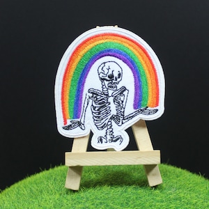 May include: Embroidered patch featuring a skeleton with a rainbow arch above it. The skeleton is depicted in a sitting position with its arms outstretched. The rainbow is in the colours of the pride flag.