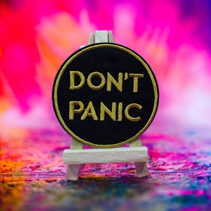May include: A black circular patch with gold embroidery that reads "DON'T PANIC" displayed on a small wooden easel against a colourful blurred background.