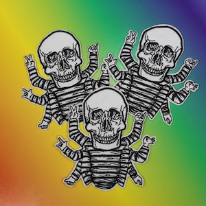 May include: Three black and white embroidered patches of skeletons with striped shirts and multiple arms in a peace sign gesture.