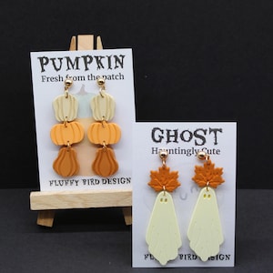 May include: Two pairs of Halloween earrings. One pair features stacked pumpkin designs in cream and orange, with the text "PUMPKIN Fresh from the patch". The other pair has ghost shapes with orange maple leaf accents, with the text "GHOST Hauntingly Cute".