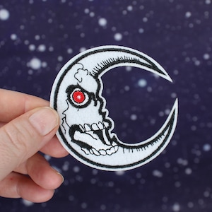 May include: A white crescent moon patch with a black skull and red eye. The patch is embroidered and has a black outline.
