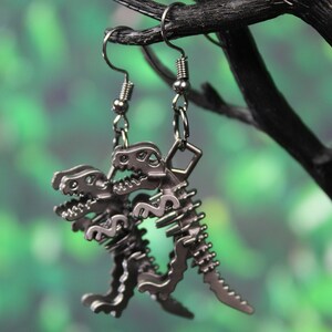 May include: A pair of silver dinosaur skeleton earrings. The earrings are hanging from a black branch against a green background.