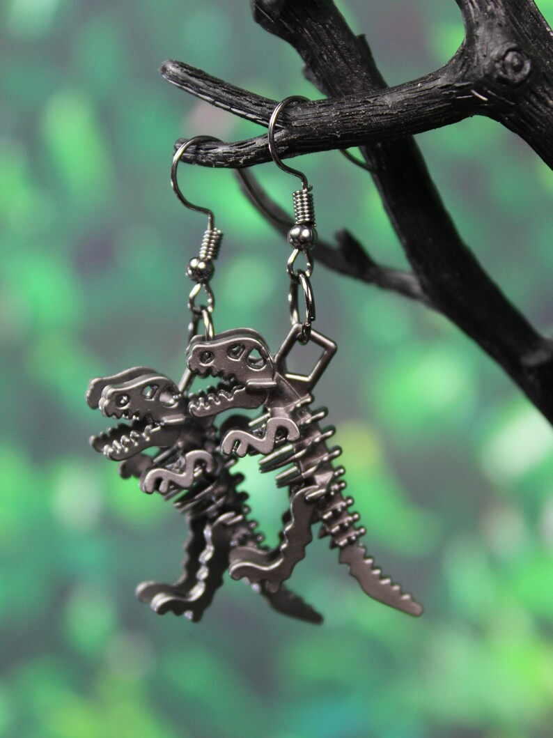 May include: A pair of silver dinosaur skeleton earrings. The earrings are hanging from a black branch against a green background.