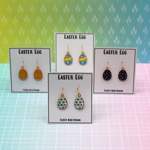 May include: Four cards with the text "EASTER EGG" and the words "FLUFFY BIRD DESIGN" on them. Each card has a pair of gold dangle earrings with different coloured and patterned Easter egg charms. The earrings are displayed on a pink and blue checkered background.