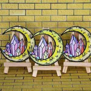 May include: Three embroidered iron-on patches featuring a crescent moon with a cluster of colorful crystals in the center. The patches are displayed on small wooden easels.