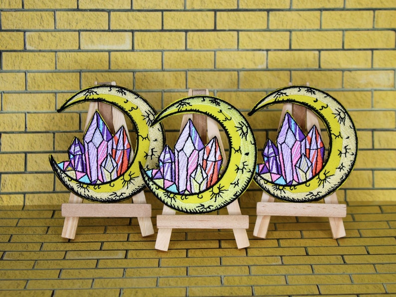 May include: Three embroidered iron-on patches featuring a crescent moon with a cluster of colorful crystals in the center. The patches are displayed on small wooden easels.