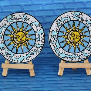 May include: Two embroidered patches featuring a sun with a face and clouds on a blue background. The patches are displayed on small wooden easels.