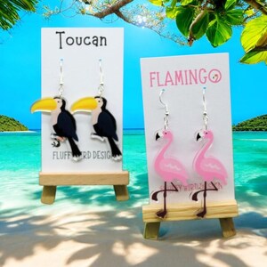 May include: Two pairs of dangle earrings featuring toucans and flamingos. The toucan earrings are black with yellow beaks and the flamingo earrings are pink. The earrings are displayed on wooden stands with white cards that say "Toucan" and "FLAMINGO".