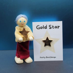 May include: A wooden doll with a yellow shirt and maroon pants holds a gold star pin. A card next to the doll reads "Gold Star" and "Fluffy Bird Design". The star pin has the text "didn't kill anyone today!"