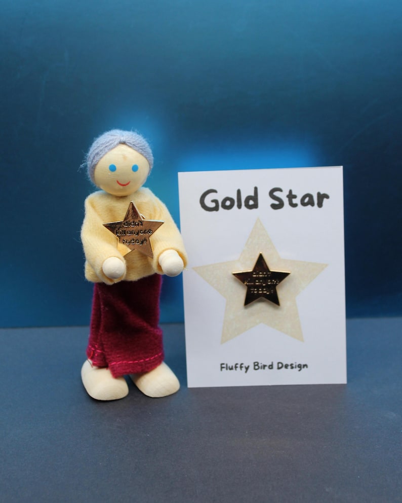 May include: A wooden doll with a yellow shirt and maroon pants holds a gold star pin. A card next to the doll reads "Gold Star" and "Fluffy Bird Design". The star pin has the text "didn't kill anyone today!"
