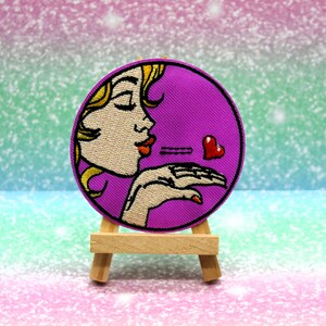 May include: Purple embroidered patch featuring a cartoon woman blowing a kiss and a red heart. The patch is displayed on a small wooden easel.