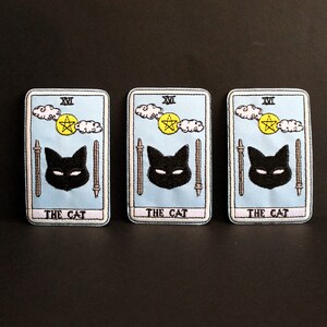 May include: Three embroidered patches with a tarot card design. Each patch features a light blue background, a black cat, a yellow pentacle, and the text "THE CAT". The patches are rectangular with a white border.