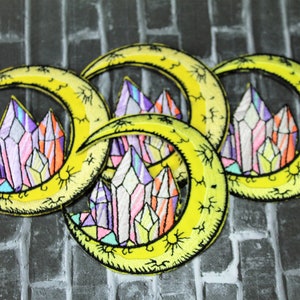 May include: Three yellow crescent moon embroidered patches with colorful crystal designs. Each patch features a crescent moon with a cluster of colorful crystals in the center. The crystals are outlined in black and have a stained glass effect.