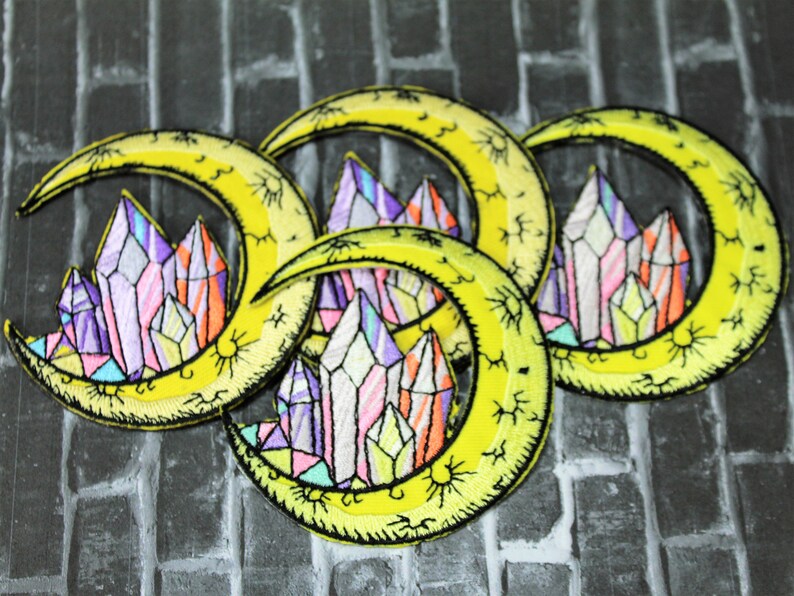 May include: Three yellow crescent moon embroidered patches with colorful crystal designs. Each patch features a crescent moon with a cluster of colorful crystals in the center. The crystals are outlined in black and have a stained glass effect.