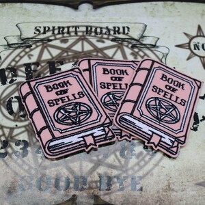 May include: Three pink fabric patches with the text "Book of Spells" and a pentagram design. The patches are in the shape of a book.
