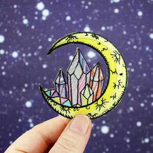May include: A yellow crescent moon with a crystal cluster inside. The moon has a detailed design with small stars and sunbursts. The crystals are multi-colored and have a rainbow effect.