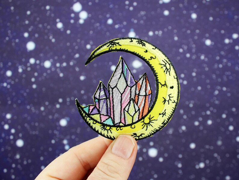 May include: A yellow crescent moon with a crystal cluster inside. The moon has a detailed design with small stars and sunbursts. The crystals are multi-colored and have a rainbow effect.