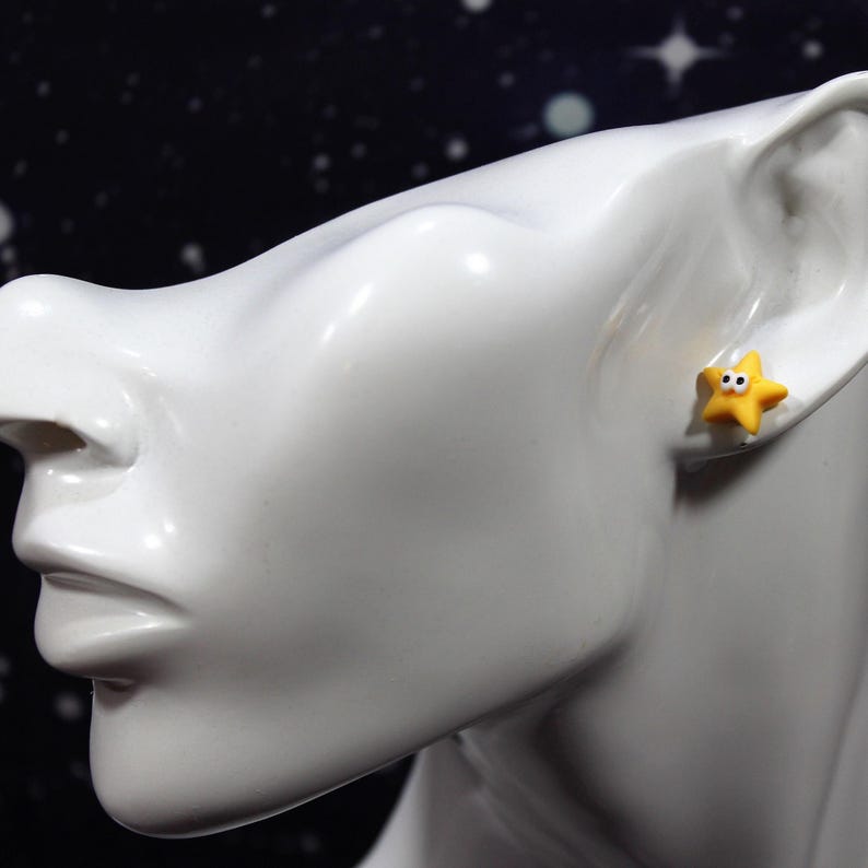 Yellow googly-eyed star earring worn on a glossy white bust, showing the fun and quirky size and fit.