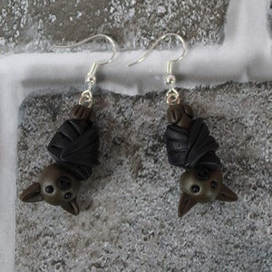 May include: A pair of dangle earrings featuring a black and brown bat design. The bats are made of polymer clay and are hanging upside down.