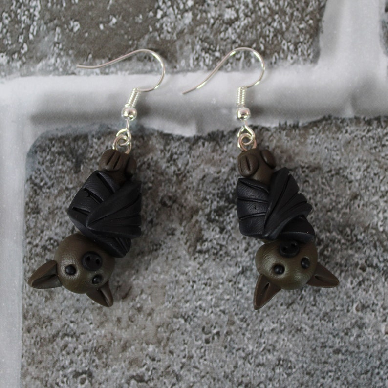 May include: A pair of dangle earrings featuring a black and brown bat design. The bats are made of polymer clay and are hanging upside down.