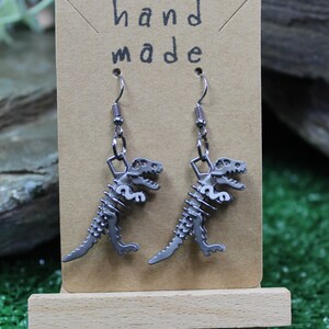 May include: A pair of silver dinosaur skeleton earrings on a brown card with the text "hand made".