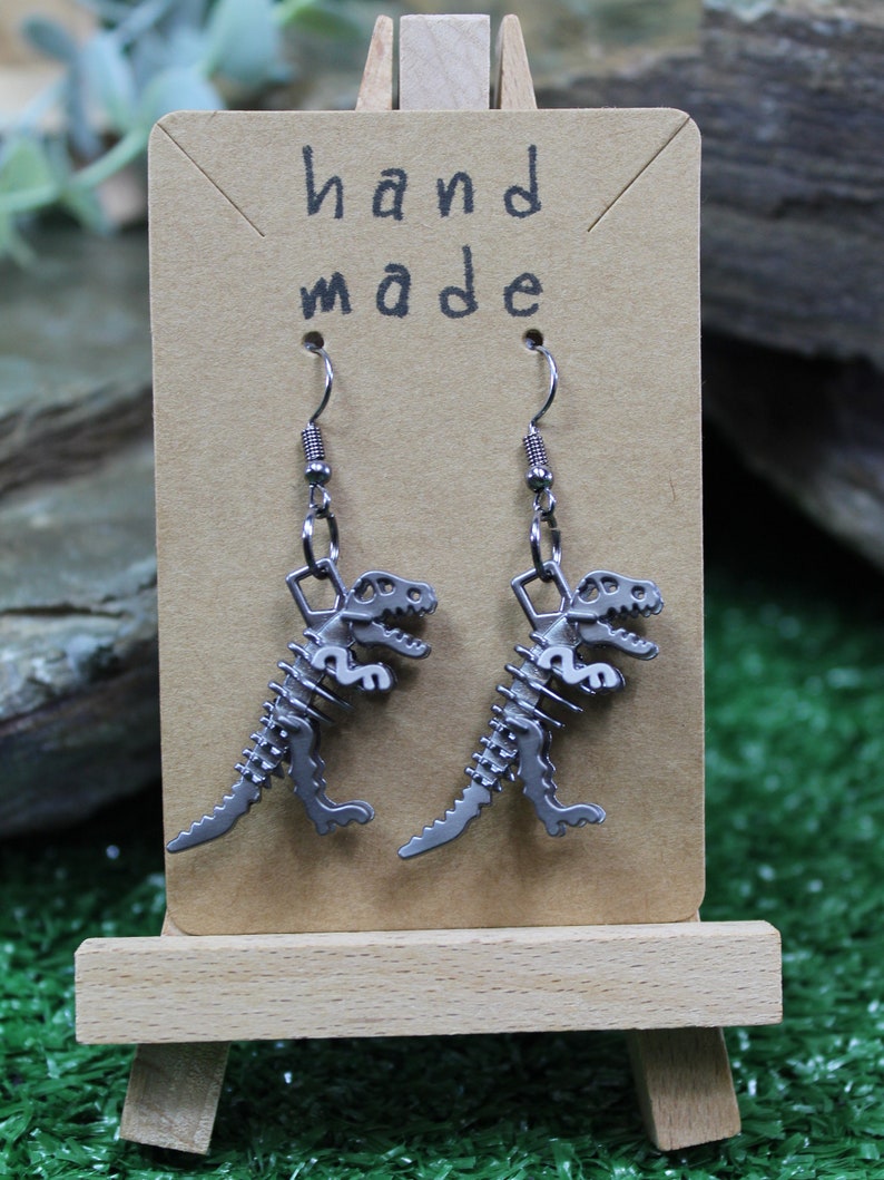 May include: A pair of silver dinosaur skeleton earrings on a brown card with the text "hand made".