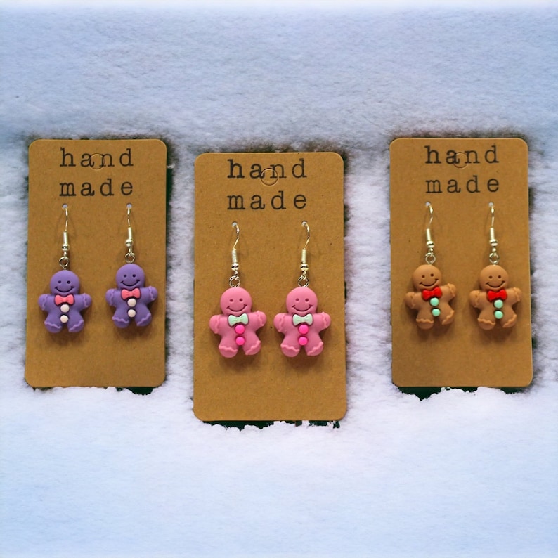 K&ouml;nnte beinhalten: Drei Paar handgemachte Lebkuchenmann-Ohrringe. Die Ohrringe befinden sich auf braunen Karten mit dem Text "hand made". Die Lebkuchenm&auml;nner sind lila, rosa und braun.