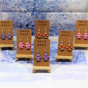 K&ouml;nnte beinhalten: Sechs Paar handgemachte Lebkuchenmann-Ohrringe, die auf kleinen Holzst&auml;ndern ausgestellt sind. Die Ohrringe sind aus Polymer Clay gefertigt und in Lila, Rosa und Braun erh&auml;ltlich.