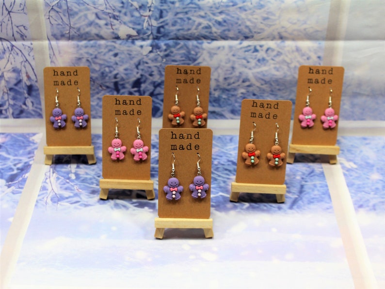 K&ouml;nnte beinhalten: Sechs Paar handgemachte Lebkuchenmann-Ohrringe, die auf kleinen Holzst&auml;ndern ausgestellt sind. Die Ohrringe sind aus Polymer Clay gefertigt und in Lila, Rosa und Braun erh&auml;ltlich.
