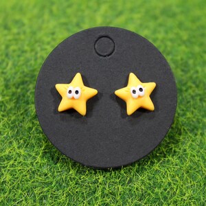 A pair of yellow star earrings mounted on a black circular card. Cartoon-style with expressive eyes, shown on faux grass. - tiny bursts of sunshine that instantly lift your mood. Picture slipping them on before you head out, and catching that cheerful star sparkle in the mirror like a little reminder to stay bright.