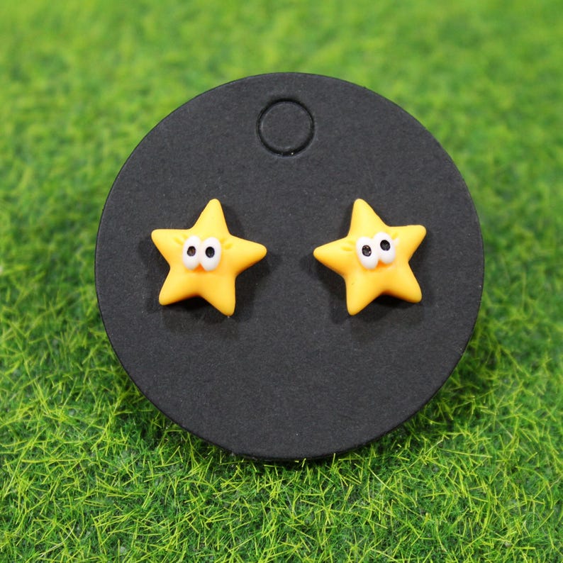 A pair of yellow star earrings mounted on a black circular card. Cartoon-style with expressive eyes, shown on faux grass. - tiny bursts of sunshine that instantly lift your mood. Picture slipping them on before you head out, and catching that cheerful star sparkle in the mirror like a little reminder to stay bright.