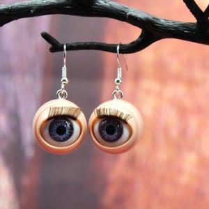 May include: A pair of earrings featuring realistic doll eyes with blue irises and brown eyelashes. The eyes are set in a flesh-toned round base and hang from silver hooks.