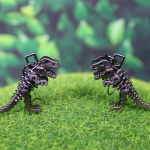 May include: Two black metal dinosaur skeleton charms, facing each other, on a green grassy surface.